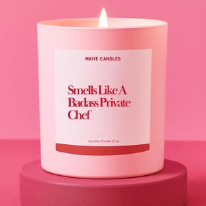 May include: A lit pink candle with a white label that reads "Smells Like A Badass Private Chef." The candle is 9 oz and sits on a matching pink pedestal against a pink background. The brand name "Maiye Candles" is at the top of the label.