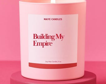 May include: A lit pink soy wax candle in a matching container, labeled "Building My Empire" with "MAIYE CANDLES" above. The candle is 9 oz and sits on a pink pedestal against a pink backdrop.