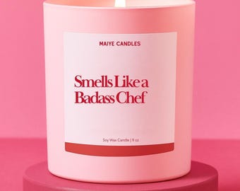 May include: A lit pink soy wax candle in a matching pink container, with the text "Smells Like a Badass Chef" on a white label. The candle sits on a round, pink pedestal. The candle is 9 oz.