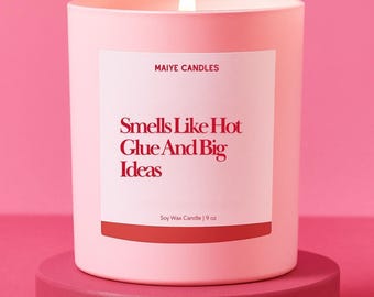 May include: A lit pink soy wax candle on a matching stand. The candle has a white label with the text "MAIYE CANDLES" and "Smells Like Hot Glue And Big Ideas." The candle is 9 oz.