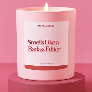 May include: A pink soy wax candle with a lit wick, labeled "Smells Like a Badass Editor." The candle is 9 oz and sits on a matching pink pedestal. The brand name "Maiye Candles" is also displayed.