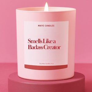 May include: A lit pink candle on a matching stand. The candle has a white label with the text "Smells Like a Badass Creator" and "MAIYE CANDLES". The candle is 9 oz.