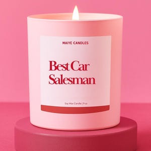 May include: A light pink candle with the text "Best Car Salesman" in red lettering. The candle is in a cylindrical container and sits on a matching pink pedestal. The candle is 9 oz.