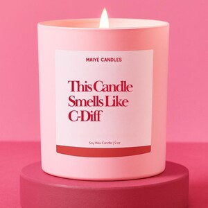 May include: A lit pink soy wax candle in a matching container, with a label that reads "This Candle Smells Like C-Diff." The candle is on a pink pedestal. The candle is 9 oz.