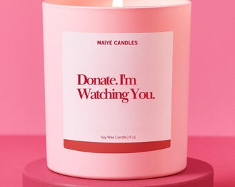 May include: A lit pink candle with the text "Donate. I'm Watching You" on a white label. The candle is on a pink stand against a pink background. The label also reads "Maiye Candles" and "Soy Wax Candle | 9 oz."