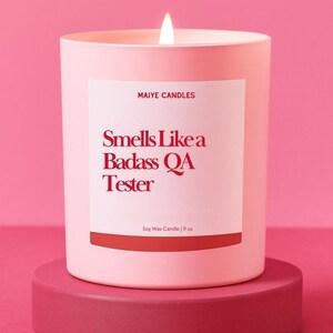 May include: A light pink soy wax candle in a matching pink jar, with a lit wick. The label reads "Smells Like a Badass QA Tester" in red text. The candle is 9 oz and sits on a pink pedestal.