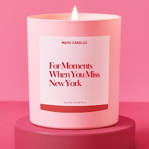 May include: A lit pink soy wax candle in a matching container, labeled "For Moments When You Miss New York." The candle sits on a round, darker pink pedestal against a pink backdrop. The label also reads "MAIYE CANDLES" and "9 oz."