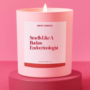 May include: A lit pink soy wax candle in a matching container, with the text "Smells Like A Badass Endocrinologist" on a white label. The candle sits on a round, pink pedestal. The candle is 9 oz.