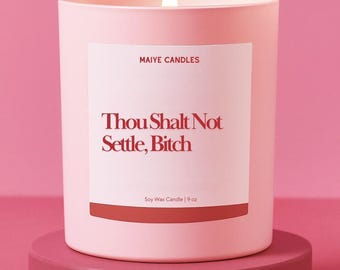 May include: A lit pink candle with the text "Thou Shalt Not Settle, Bitch" on a white label. The candle is 9 oz and sits on a matching pink stand. The brand name "Maiye Candles" is above the text.