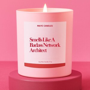 May include: A lit pink soy wax candle in a matching container, with a white label that reads "Smells Like A Badass Network Architect." The candle sits on a round, pink pedestal. The candle is 9 oz.