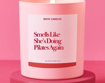 May include: A lit pink candle with the text "Smells Like She's Doing Pilates Again" on a white label. The candle is 9 oz and sits on a matching pink pedestal against a pink background. The brand name "MAIYE CANDLES" is at the top.