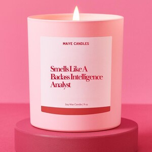 May include: A lit pink soy wax candle in a matching container, labeled "Smells Like A Badass Intelligence Analyst." The candle sits on a round, pink pedestal. The candle is 9 oz.