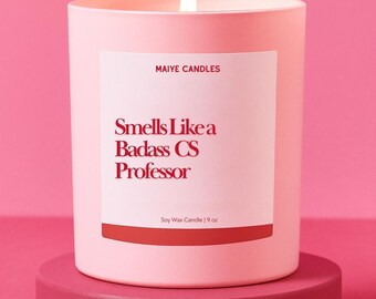 May include: A lit pink candle with the text "Smells Like a Badass CS Professor" on a white label. The candle is 9 oz and sits on a matching pink pedestal, against a pink background. The brand name "Maiye Candles" is also visible.