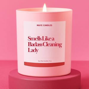 May include: A lit pink soy wax candle in a matching pink container, with the text "Smells Like a Badass Cleaning Lady" on a white label. The candle sits on a pink pedestal against a pink background. The candle is 9 oz.