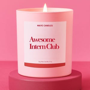 May include: A lit pink soy wax candle in a matching container, labeled "Awesome Intern Club" in red text. The candle is 9 oz and sits on a pink pedestal against a pink background. The brand name "Maiye Candles" is also visible.