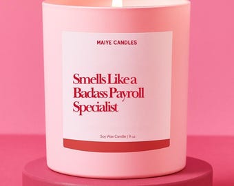 Puede incluir: Una vela rosa encendida de cera de soja en un recipiente a juego, con el texto "Smells Like a Badass Payroll Specialist". La vela es de 255 gramos y se encuentra sobre un pedestal rosa, sobre un fondo rosa.