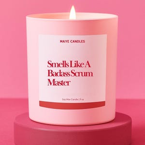 May include: A lit pink soy wax candle in a matching container, labeled "Smells Like A Badass Scrum Master." The candle sits on a pink pedestal. The label also reads "Maiye Candles" and "9 oz."