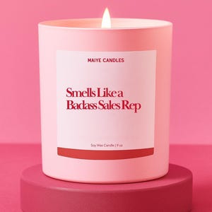 May include: A lit pink candle in a matching container sits on a pink pedestal. The candle has a white label with the text "Smells Like a Badass Sales Rep" in red. The label also reads "MAIYE CANDLES" and "Soy Wax Candle | 9 oz."