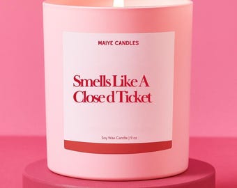 May include: A lit pink soy wax candle in a matching container, labeled "Smells Like A Closed Ticket" by Maiye Candles. The candle sits on a round, pink pedestal. The candle is 9 oz.