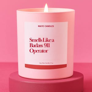 May include: A lit pink candle with the text "Smells Like a Badass 911 Operator" on a white label. The candle is on a matching pink stand. The candle is 9 oz.