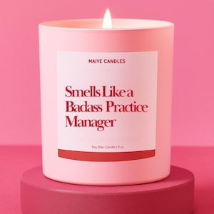 May include: A lit pink soy wax candle on a matching pink pedestal. The candle has a white label with the text "Smells Like a Badass Practice Manager" in red. The candle is 9 oz.