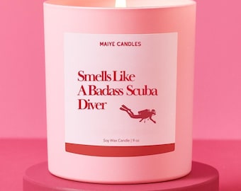 May include: A pink soy wax candle with a lit wick. The label reads "Smells Like A Badass Scuba Diver" with a diver graphic. The candle is 9 oz and sits on a matching pink stand. The brand name "Maiye Candles" is at the top.