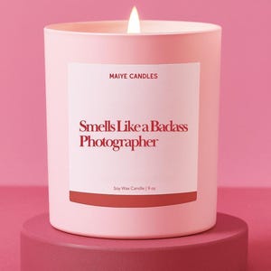 May include: A lit pink soy wax candle with the text "Smells Like a Badass Photographer" on a white label. The candle is 9 oz and sits on a matching pink pedestal. The brand name "Maiye Candles" is also on the label.