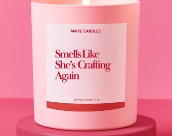May include: A lit pink candle on a matching pink stand. The candle has a white label with the text "Smells Like She's Crafting Again" and "Soy Wax Candle | 9 oz". The candle is from "Maiye Candles".