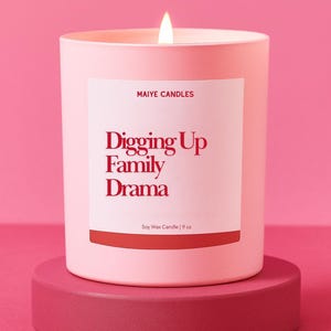 May include: A light pink soy wax candle in a matching pink container, with a white label featuring the text "Digging Up Family Drama" in red. The candle is 9 oz and sits on a pink pedestal, with a lit wick.