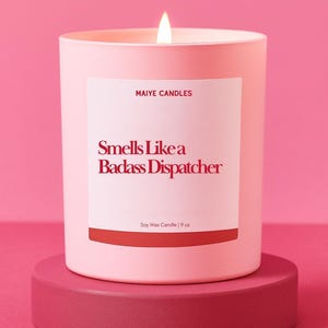 May include: A pink candle with a lit wick, labeled "Smells Like a Badass Dispatcher." The candle is in a pink container and sits on a matching pink pedestal. The label also reads "Maiye Candles" and "Soy Wax Candle | 9 oz."
