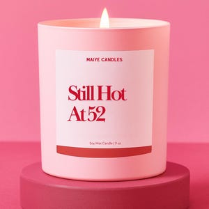 May include: A lit pink soy wax candle in a matching pink container, with the text "Still Hot At 52" in red. The candle sits on a round, dark pink pedestal against a pink background. The label also reads "Maiye Candles" and "Soy Wax Candle | 9 oz."