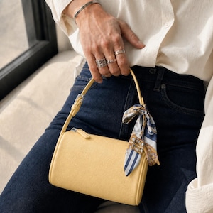 May include: A cylindrical yellow handbag with a single adjustable strap and a silver zipper closure. A decorative silk scarf featuring a blue, white, and gold patterned print is tied around the base of the strap, adding a distinct visual accent.