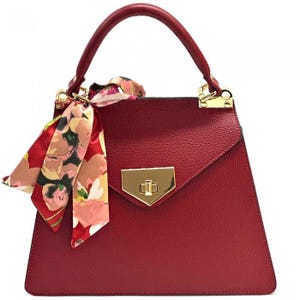 May include: A red handbag with a top handle and a decorative floral scarf tied to the handle. The bag has a gold-colored clasp and hardware. The bag's shape is trapezoidal.