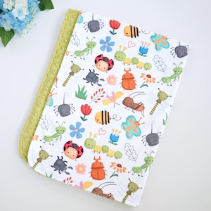 May include: A white baby blanket with a colorful insect and floral pattern. The blanket features various cartoon insects, flowers, and leaves. The border is a textured, light green fabric.