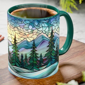 May include: A ceramic mug with a stained-glass mountain scene in shades of blue, green, and yellow. The mug has a dark green handle and rim, and is filled with a dark liquid. The mug is on a wooden cutting board.