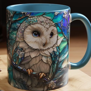 Op de afbeelding: Een keramische koffiemok met een blauw handvat en interieur. De mok heeft een gedetailleerde illustratie van een uil met blauwe ogen, zittend op een tak, tegen een achtergrond in glas-in-stijl met tinten blauw en groen.