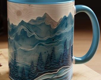 May include: A ceramic mug with a blue interior and handle, featuring a mountain and forest design in shades of blue and white. The mug is set on a wooden surface with a spoon, coffee beans, and a pastry.