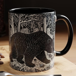 May include: Black and white ceramic mug depicting a bear in a forest scene. The mug has a black interior and handle. The design shows detailed trees and foliage. A spoon and a pastry are on a wooden surface.