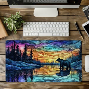 Puede incluir: Alfombrilla de ratón rectangular con un paisaje colorido. Representa un oso de pie en un lago, con una puesta de sol, árboles y montañas. La paleta de colores incluye azules, naranjas y morados.