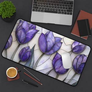 Pode incluir: Tapete de rato retangular com um design de vitral de tulipas roxas e folhas verdes sobre um fundo branco. Um laptop, café e artigos de papelaria são visíveis.