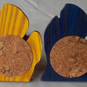 May include: Two wooden coasters, one yellow and one blue, each shaped like a stylized hand. A round cork coaster is affixed to the front of each wooden piece. The coasters are designed to protect surfaces from drink condensation.