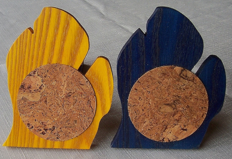May include: Two wooden coasters, one yellow and one blue, each shaped like a stylized hand. A round cork coaster is affixed to the front of each wooden piece. The coasters are designed to protect surfaces from drink condensation.