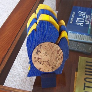 May include: A wooden sculpture with a blue and yellow layered design, featuring a circular cork element in the center. The sculpture is mounted on a small blue base, showcasing a unique artistic style.