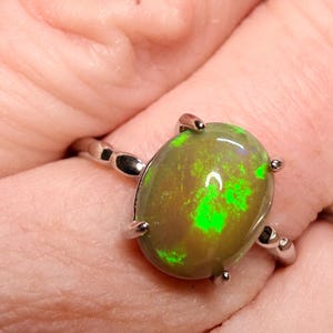 May include: A silver ring featuring an oval-shaped opal gemstone. The opal has a dark base color with vibrant green flashes. The ring's band is thin and delicate, with a simple design. The gemstone is held in place by four prongs.