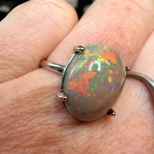 May include: A silver ring featuring a large, oval opal gemstone. The opal displays a vibrant play of color, with flashes of red, green, and yellow. The gemstone is held in place by four small prongs.