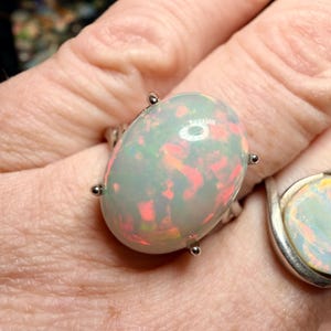 May include: Oval-shaped opal ring with a silver band. The opal has a milky white base with flashes of red and green. Four silver prongs secure the opal to the band. Another ring with an opal is visible in the background.