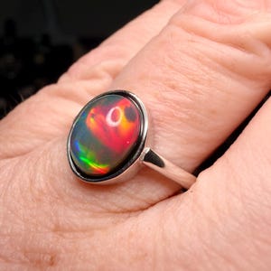 May include: A silver ring featuring a large, oval-shaped black opal cabochon. The gemstone displays vibrant flashes of red, orange, and green play-of-color across its dark surface. The stone is set in a smooth, polished silver bezel on a simple band.