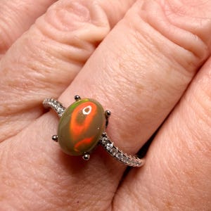 May include: A silver-toned ring featuring a central oval-shaped cabochon opal with vibrant orange and green flashes of color. The band is set with small, clear, sparkling stones along the sides and is held in place by four metal prongs.