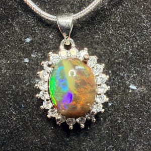 May include: A silver pendant necklace featuring an oval opal gemstone with a vibrant play of color. The opal is surrounded by a halo of small, clear gemstones, and hangs from a delicate silver chain.