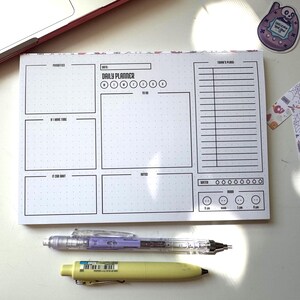 Op de afbeelding: Een witte dagelijkse planner-notitieblok met een zwarte rasterindeling, met secties voor prioriteiten, takenlijsten en notities. Een doorzichtige mechanische potlood met paarse accenten en een gele pen liggen naast het notitieblok. Een sticker en decoratieve tape zijn ook aanwezig.